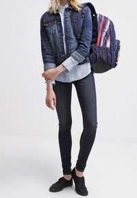 Joven con una chaqueta de mezclilla, camisa estampada, jeans ajustados, zapatillas negras y llevando una mochila a rayas colorida en un hombro.