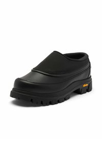 Schwarzer Slip-On-Schuh mit dicker, strukturiertem Vibram-Sohle, gepolstertem Kragen und glattem Obermaterial mit Nahtdetails auf weißem Hintergrund.