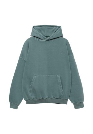 Person trägt einen türkisfarbenen Hoodie mit kleinem besticktem Text auf dem Rücken über einem braun karierten Hemd und dunklen Denim-Jeans, von hinten gesehen.