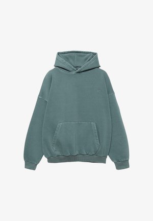 Personne portant un sweat à capuche bleu sarcelle avec un petit texte brodé dans le dos, par-dessus une chemise à carreaux marron et un jean en denim foncé, vue de dos.