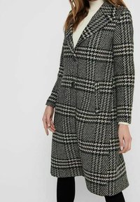 Schwarz-weißer karierter Mantel mit Kragen, zwei Knöpfen und seitlichen Taschen. Texturiertes Gewebe und knielanges Design. Über einem weißen Oberteil getragen.