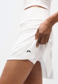 Λευκή αθλητική φούστα από λείο ύφασμα, με μαύρο λογότυπο στο τελείωμα και σχεδιασμό με στρώσεις για μεγαλύτερη κίνηση.