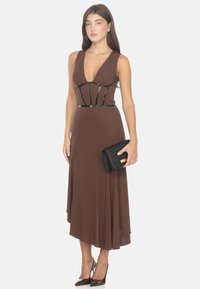 Abito marrone senza maniche con scollatura a V profonda e dettagli stile corsetto in vita; abbinato a una pochette nera e scarpe con tacco alto.