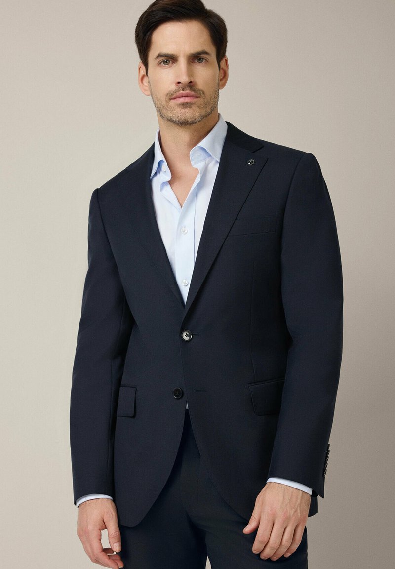 Granatowa marynarka dopasowana, z dwoma guzikami, klapami w kształcie notcha i kieszeniami z przodu. Wykonana z teksturowanej tkaniny, zestawiona z jasnoniebieską koszulą z kołnierzykiem.