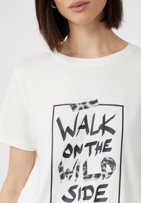 T-shirt en coton blanc avec un col rond, présentant un imprimé graphique noir audacieux et des accents de motif léopard dans le texte.