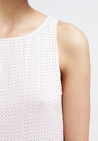 Ärmlös topp i ljusrosa tyg med ett mönster som har små mörkblå former. Slät textur med rund halsringning.