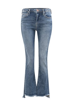 Kobieta stojąca w białym t-shircie z grafiką, niebieskich skróconych dżinsach z postrzępionymi krawędziami oraz brązowych sandałach na jednolitym szarym tle.