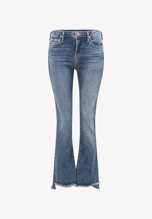 Vrouw staand, draagt een wit shirt met print, blauwe korte spijkerbroek met rafelige zomen en bruine sandalen tegen een effen grijze achtergrond.