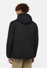 Mustahuppari, jossa on vettä hylkivä pinta, joustavat hihat ja yksinkertainen muotoilu. Takana on sileä pinta eikä näkyvää logoa.