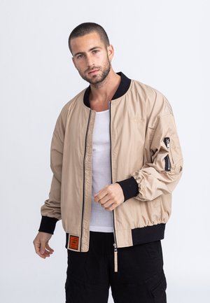 Beige Bomberjacke mit schwarzem Rippkragen und -bündchen, Reißverschluss vorne und aufgesetzter Tasche am linken Ärmel. Verfügt über eine strukturierte Außenseite.