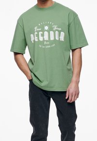 Green cotton t-shirt with short sleeves and round neck. Features front graphic text: "WELCOME Finer Things PEGADOR Italy TO THE GOOD LIFE."