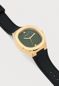 Goldfarbene Guess-Armbanduhr mit grün strukturiertem Zifferblatt, schwarzem Silikonarmband und minimalistischen Stundenmarkierungen bei 12 und 6 Uhr.