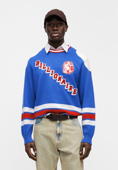 Μπλε πλεκτό πουλόβερ με κόκκινες και λευκές ρίγες, κείμενο "BILLIONAIRE" και γραφικό αστροναύτη. Συνδυασμένο με τζιν παντελόνι ανοιχτού τόνου και καφέ καπέλο.