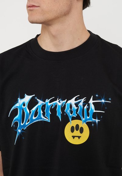 Young man wearing a black T-shirt with shiny blue graffiti-style text and a yellow fanged paw print graphic.