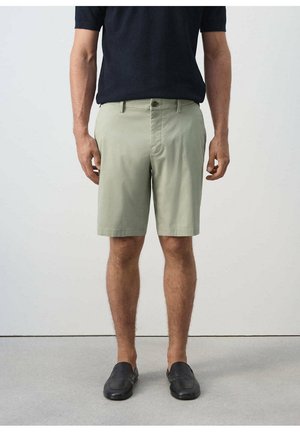 Hombre con pantalones cortos hasta la rodilla de color verde claro, zapatos negros sin cordones y una camisa oscura de manga corta, de pie sobre un fondo liso.