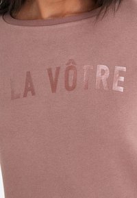 Bleget lyserød sweatshirt med rund hals og korte ærmer, med skinnende tekst "LA VÔTRE" i en kontrastfarve foran.
