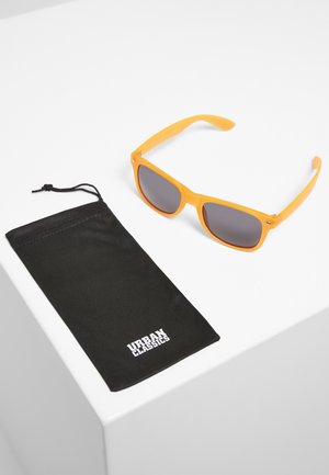 Oransje plastsolbriller med mørke linser ved siden av en svart myk pose, trykt med "URBAN CLASSICS" i hvitt på forsiden.
