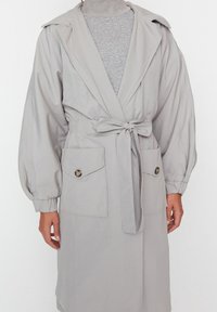Manteau portefeuille gris clair en tissu lisse avec une taille ceinturée, des manches surdimensionnées et deux poches avant fermées par des boutons.