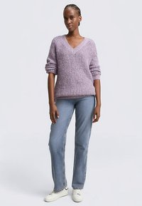 Lavendelfarbener Strickpullover mit V-Ausschnitt und hochgekrempelten Ärmeln, kombiniert mit hellblauen Jeans und weißen Sneakern. Strukturierter, lockerer Sitz.