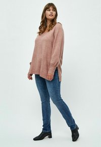 Hellrosa, übergroßer Strickpullover mit V-Ausschnitt und gerippten Bündchen, kombiniert mit blauen engen Jeans und schwarzen, gefütterten Ankle-Boots mit Absatz.