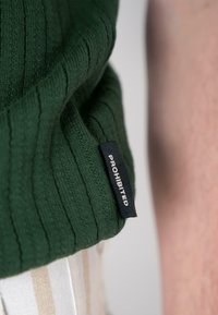 Green ribbed knit garment with a tag reading "PROHIBITED." The garment has a textured surface and is partially visible near a striped fabric.