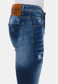 Denim jeans in een vervaagde blauwe wassingen met gescheurde details, een leren label met de tekst "BLACK BULL," en vijf-pocket stijl.
