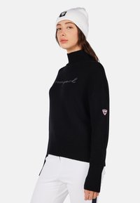Maglione a collo alto in maglia nera con orlo a costine, dotato di logo ricamato sul petto e un'etichetta con logo sulla manica sinistra. Indossato con pantaloni bianchi.