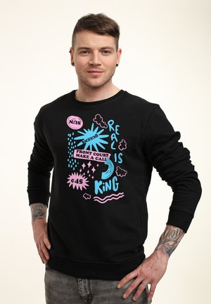 Jeune homme aux cheveux courts et tatouages sur les avant-bras porte un sweat-shirt noir avec des textes et graphiques colorés, mains sur les hanches.