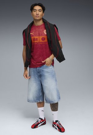 Jersey rosso a fantasia con logo, abbinato a pantaloni corti di jeans blu, giacca nera drappeggiata sulle spalle e sneakers rosse e nere.