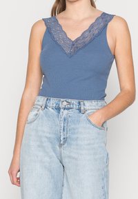 Blå ribbad tanktopp med spetsprydd V-ringning. Bärs med ljus tvättade högmidjade jeans. Tätt sittande design som framhäver den mjuka texturen.
