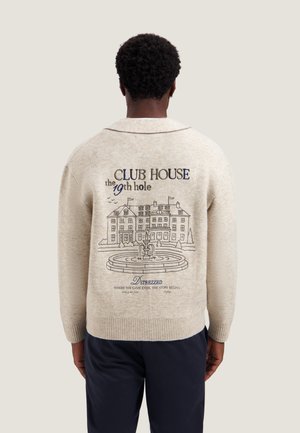 Personne portant un pull beige avec broderie d'un club-house et d'une fontaine et texte au dos, debout devant un fond uni.