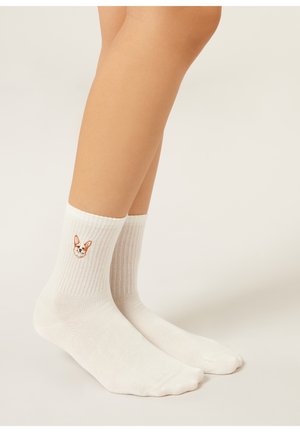 Bílé žebrované ponožky s malou vyšívanou tváří psa corgi, nošené na nohou na jednobarevném světle pozadí.