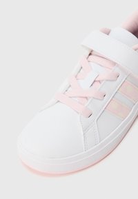 Bílá teniska s růžovými akcenty, s hladkým koženým svrškem, šněrováním a puntíkovaným vzorem na boku. Zaoblená špička a gumová podrážka.