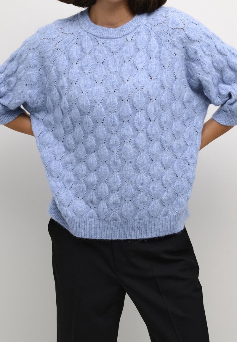 Personne portant un pull en tricot texturé bleu clair à manches trois-quarts et un pantalon noir, les mains posées sur le bas du dos.