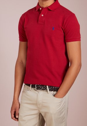Homme portant un polo rouge à manches courtes avec un logo bleu brodé et un pantalon beige avec une ceinture tressée marron.