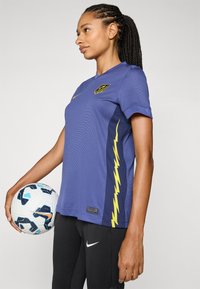 Maglietta sportiva viola con collo a giro, caratterizzata da una striscia gialla a forma di fulmine lungo il lato, abbinata a pantaloni sportivi neri e a un pallone da calcio.