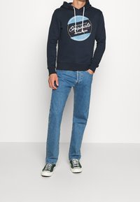 Marinblå huvtröja med en grafisk logotyp, kombinerad med ljusblå raka jeans och gröna sneakers med vita sulor.