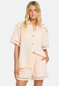 Camicia rosa chiaro a maniche corte con chiusura a bottoni e dettagli ricamati. Abbinata a pantaloni corti coordinati con tessuto testurizzato e tasche.