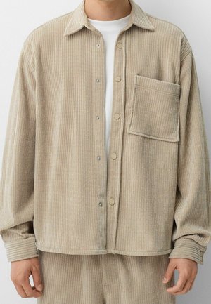 Veste en velours côtelé beige à côtes avec grande poche poitrine, boutons-pression, portée sur une chemise blanche par une personne montrée du cou aux hanches.