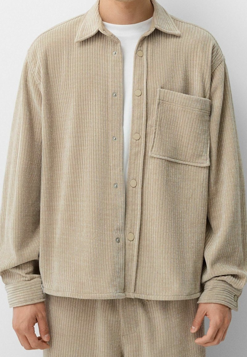 Veste en velours côtelé beige à côtes avec grande poche poitrine, boutons-pression, portée sur une chemise blanche par une personne montrée du cou aux hanches.