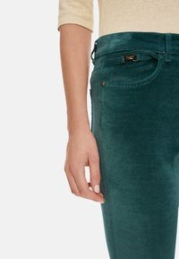 Teal cotton corduroy pants featuring a fitted design, belt loops, side pockets, and a smooth texture. Includes a subtle metallic accent.