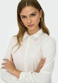 Blouse blanche à manches longues avec un col décoratif gris clair et des boutons sur le devant. Tissu doux avec une légère brillance.