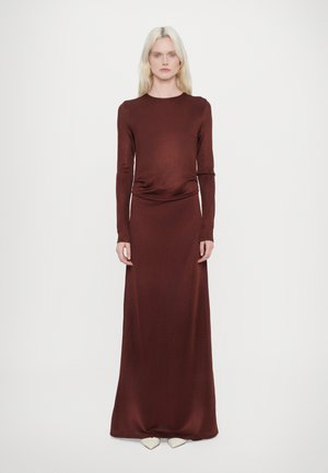 Rochie burgundy cu mânecă lungă, cu un corset ajustat și o fustă fluidă, având o talie plisată și o textură netedă. Pantofi albi ascuțiți vizibili.