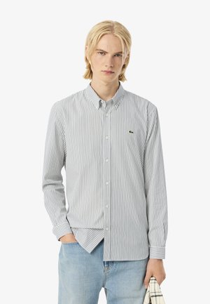 Joven con cabello largo y rubio, vistiendo una camisa de botones a rayas y pantalones jeans de color azul claro, sosteniendo un paño doblado en una mano.