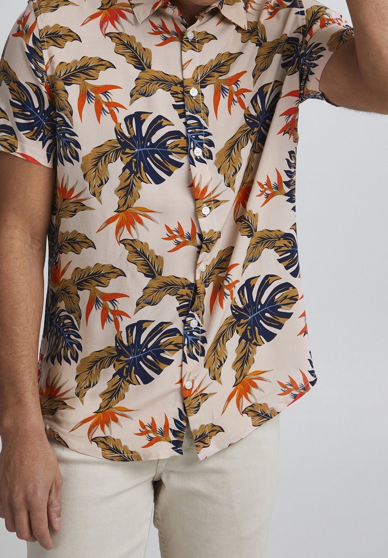 Homme portant une chemise beige à manches courtes boutonnée avec un motif de feuilles tropicales bleu marine, marron et orange, et un pantalon clair.