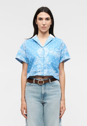 Γυναίκα με μακριά σκούρα μαλλιά που φοράει ένα μπλε φλοράλ κοντομάνικο crop top με κουμπιά και ανοιχτό μπλε τζιν με καφέ ζώνη.