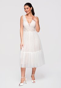 Femme souriante, portant une robe blanche en dentelle sans manches avec un profond décolleté en V et une jupe midi transparente à pois, associée à des talons blancs à bride cheville.