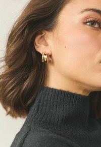 Zelta, skulpturālas auskari ar viļņotu dizainu. Modelis valkā melnu ribotā adījumā džemperi ar augstu apkakli. Mati ir vidēja garuma un viļņaini.