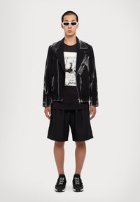 Bărbat purtând ochelari de soare albi, jachetă din piele albă, tricou grafic negru, pantaloni scurți negri și adidași negri cu text alb.