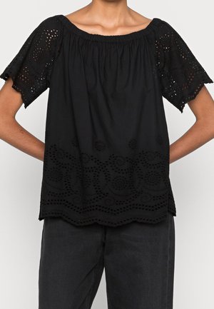 Personne portant un chemisier noir à manches courtes avec broderie œillets sur les manches et l'ourlet, associé à un pantalon noir, mains sur les hanches.